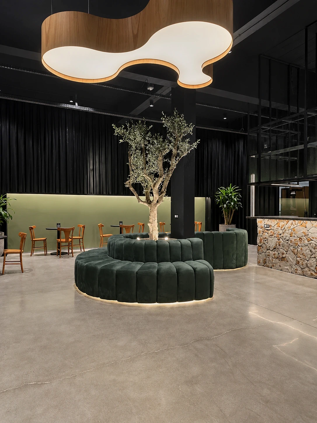 Luminária orgânica, oliveira central e banco circular no salão.